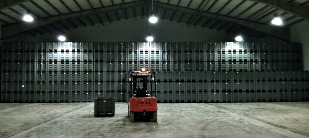 in a large warehouse, there are many stacked box pallets and in fork-lift is in front of all of them 