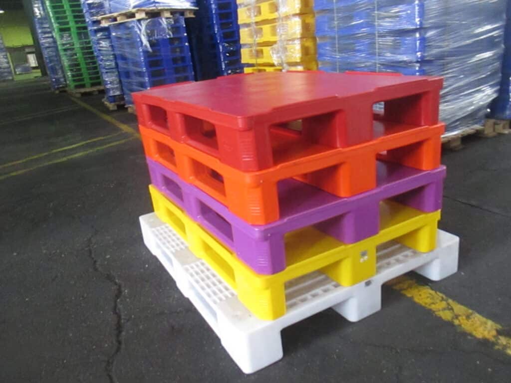 the photos describe a pile of five plastic pallet made of HDPE injection grade material in red, orange, purple, yellow and white colors on the floor of a warehouse. The Plastic Pallets are produced based on HDPE material & the injection molding method